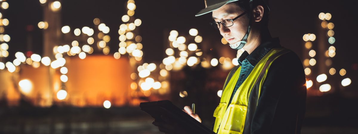 Asian man engineer using digital tablet working late night shift at petroleum oil refinery in industrial estate. Chemical engineering, fuel and power generation, petrochemical factory industry concept